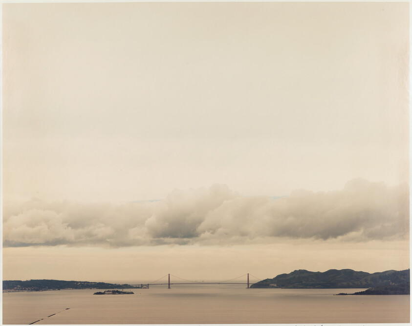 Golden Gate Bridge spans calm bay between hills under a wide, cloudy sky.