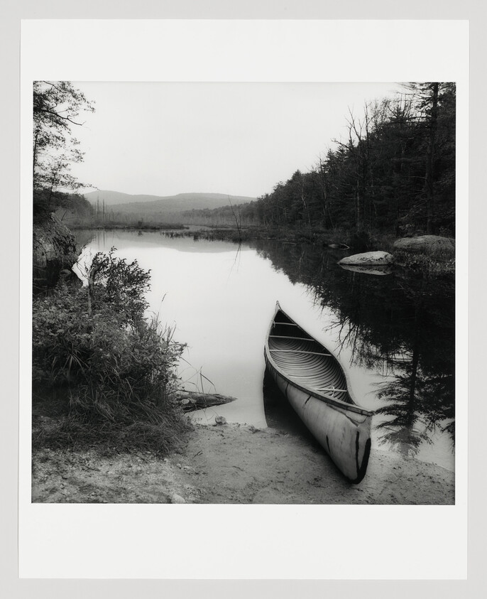 A canoe rests at the calm lakeshore with trees reflecting on the water.