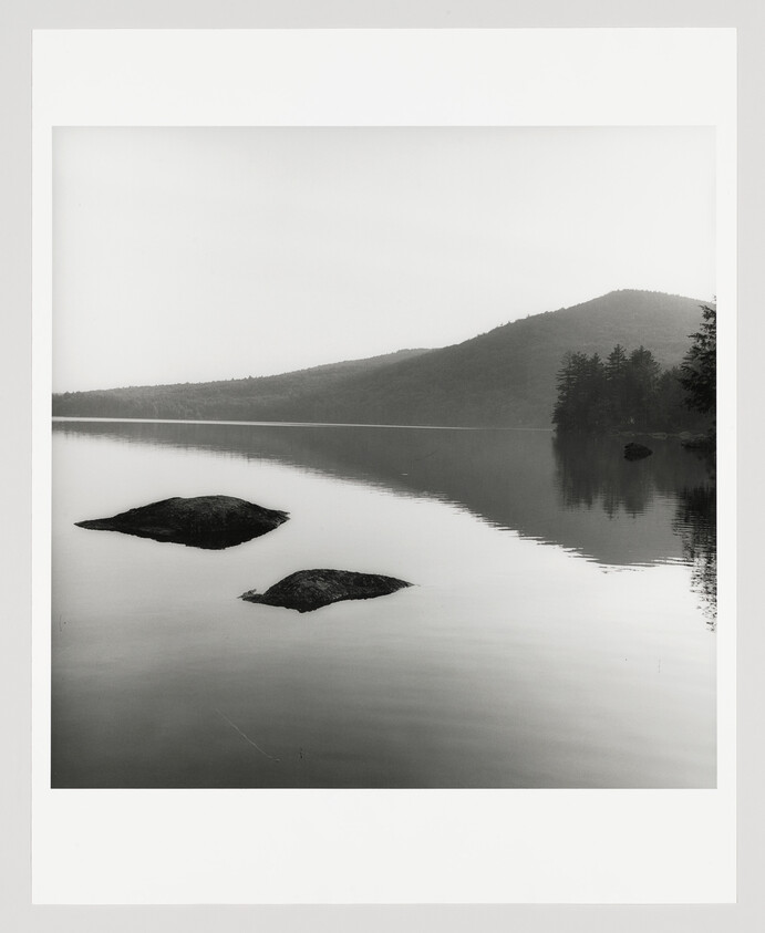 Two partially submerged rocks sit in a calm lake reflecting a distant tree-covered hill.