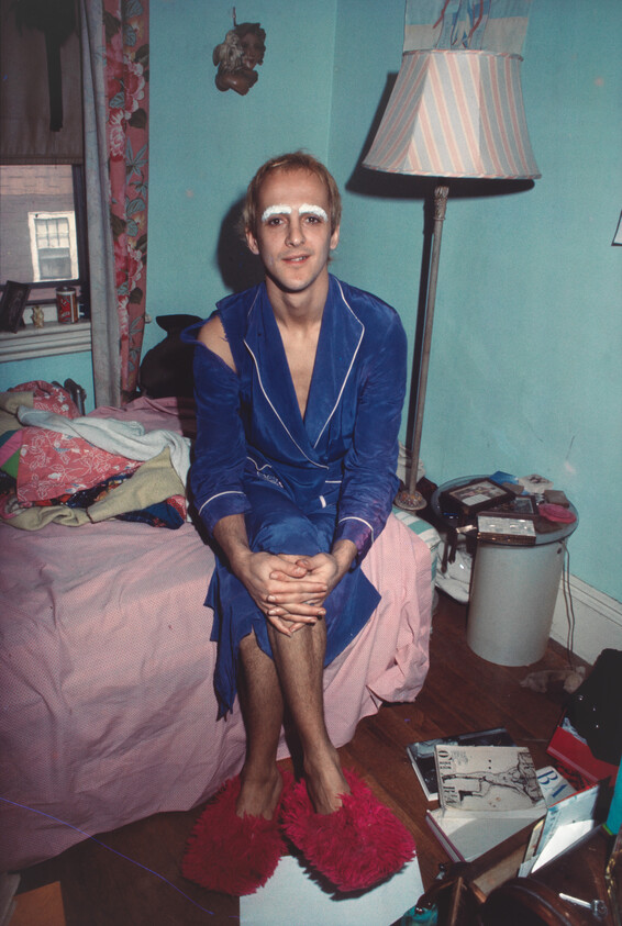 Man in blue robe with white eyebrow makeup sits on bed wearing red fuzzy slippers.