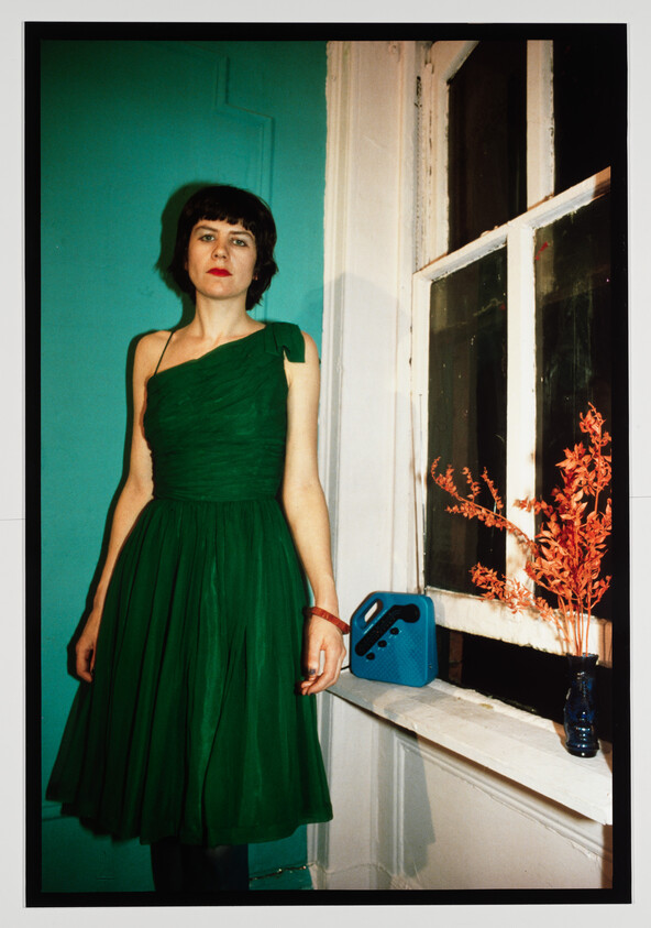 A woman in a green dress stands by a window with orange flowers and a blue radio.