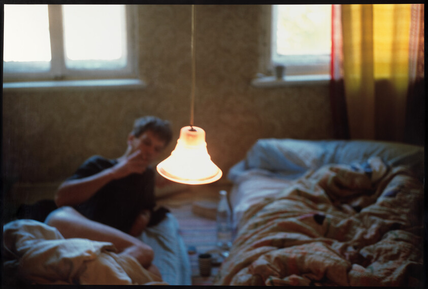A dimly lit room with a glowing hanging lamp and a person sitting on a rumpled bed smoking.