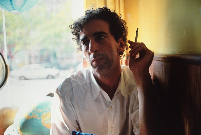 A man sits by a window holding a cigarette and looking thoughtful beside a globe.