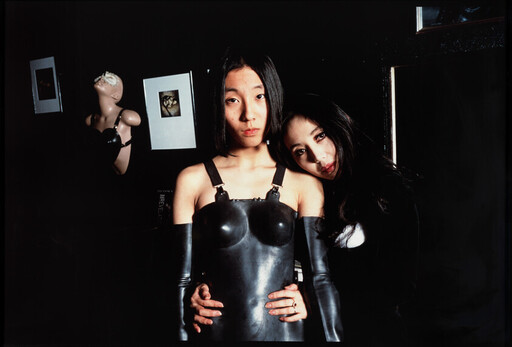Two women pose in a dark room, one wearing a black latex dress while the other leans and holds her waist.