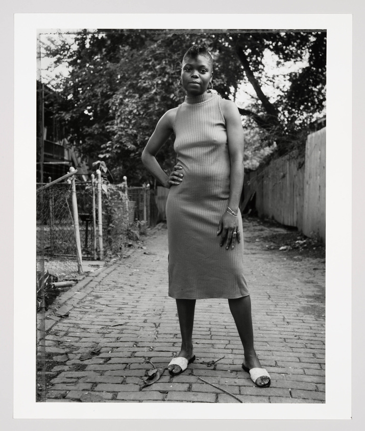 In black and white, a young Black woman stands on a fenced brick street with one hand on her hip looking straight into the camera. 