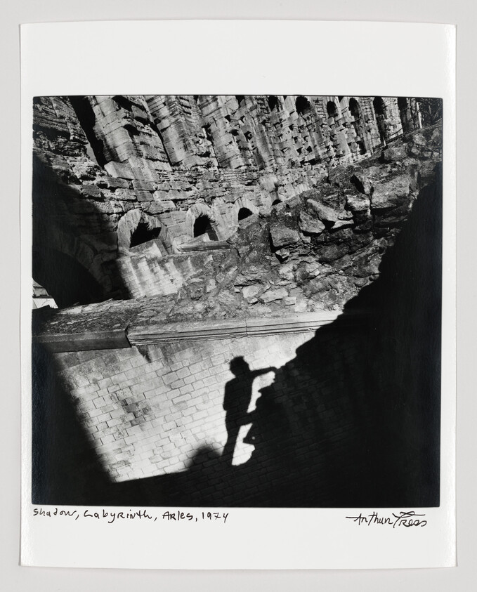 A person's shadow climbs a stone wall beside ancient ruined arches and rocky terraces.