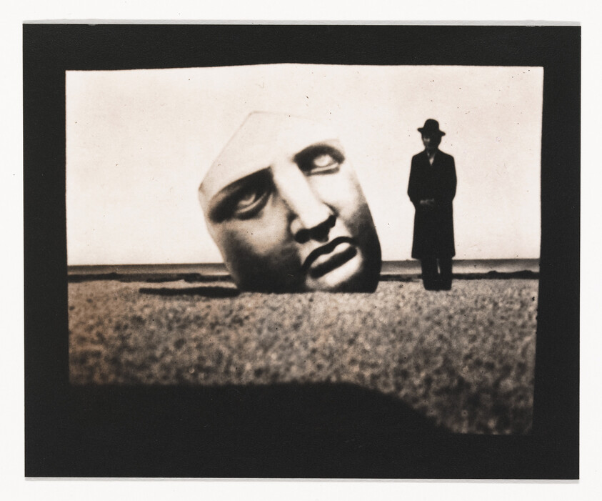 A large sculpted head lies on a beach while a man in a coat and hat stands nearby.