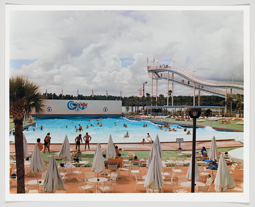 A bustling water park scene with people swimming and lounging around a large pool. In the background, there's a prominent "Wet'n Wild" sign on a wall, and a tall water slide structure extends into the sky. The foreground shows sun loungers and folded umbrellas on a tiled deck, with a few palm trees adding to the tropical ambiance. The sky is partly cloudy, suggesting a warm but overcast day.
