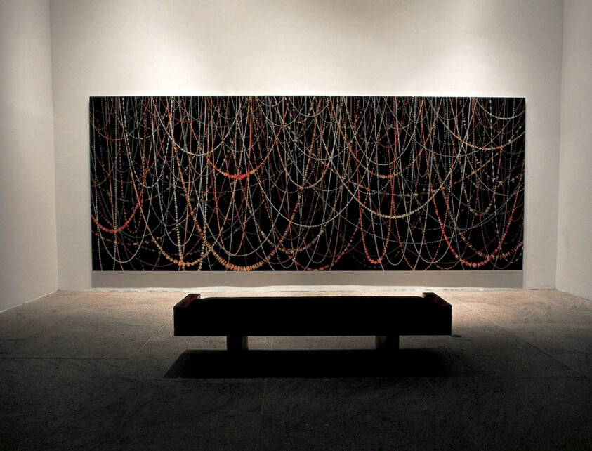Large dark painting with cascading beaded strings hangs above a low bench in a dim gallery.