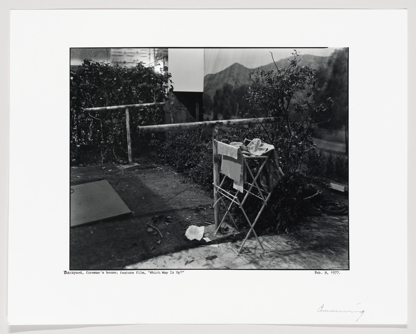Black and white photograph depicting a staged outdoor scene with a wooden fence, laundry on a folding rack, and a backdrop showing a mountain landscape. The image includes text at the bottom indicating it's a backyard from a foreman's house in a feature film, dated February 8, 1977, with a signature on the bottom right.