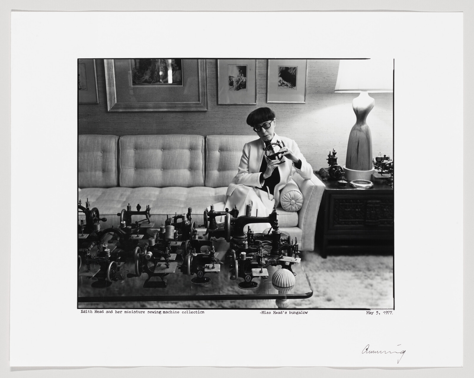 A black and white photograph depicting a woman sitting on a sofa, examining a miniature sewing machine from a collection displayed on a coffee table in front of her. The room is elegantly furnished with a cushioned sofa, framed artwork on the wall, and a mannequin with a dress in the background. The photo includes a caption at the bottom indicating the woman's name and the collection, as well as the location and date of the photograph.