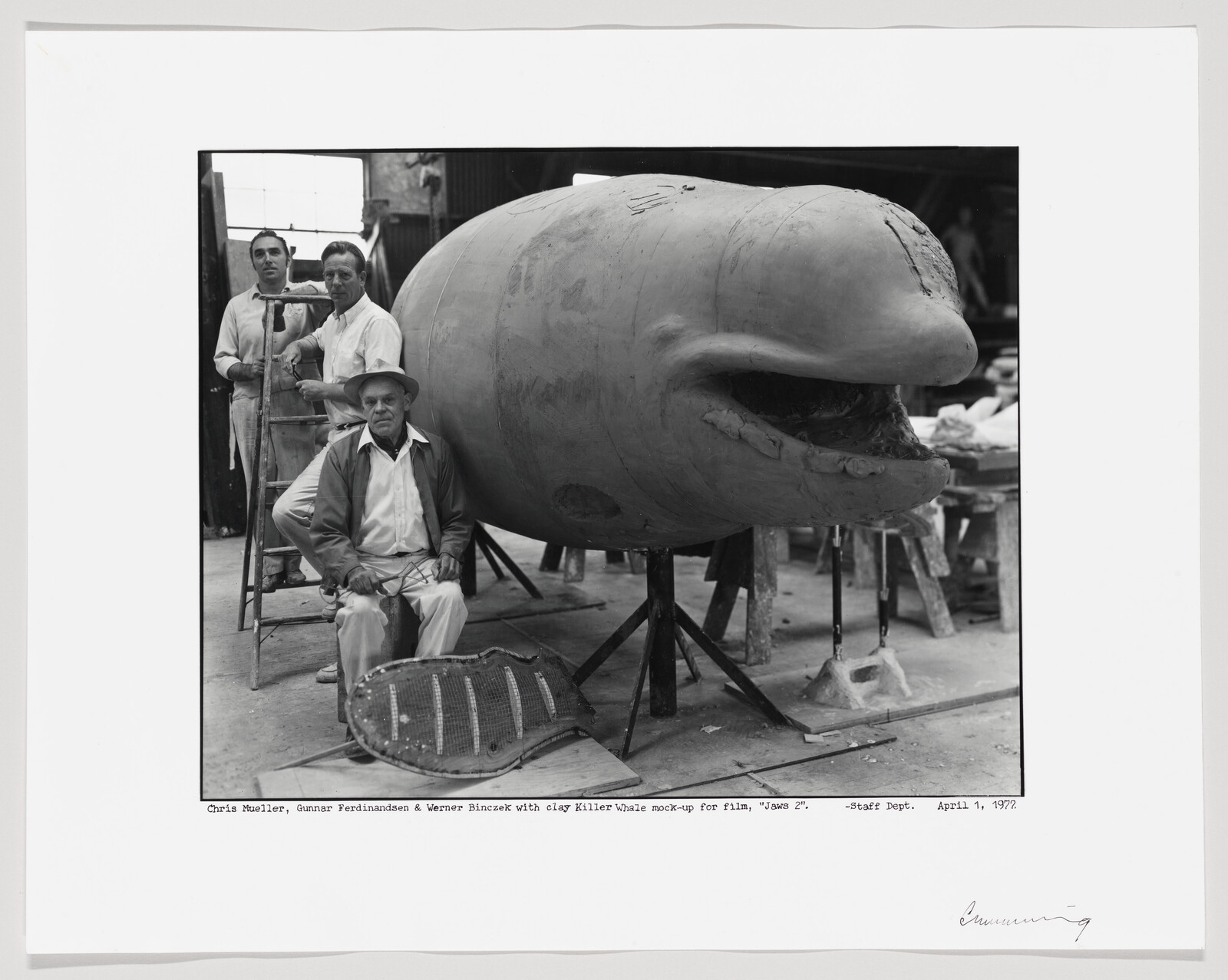 A black and white photo showing three men in a workshop with a large clay model of a killer whale. One man is seated in front of the model, while the other two stand behind it, one on a ladder. The model is on a stand and appears to be a work in progress. There is handwriting and typed text at the bottom providing details about the individuals and the purpose of the model, which is for a film titled "Jaws 2". The date indicated is April 1, 1977.