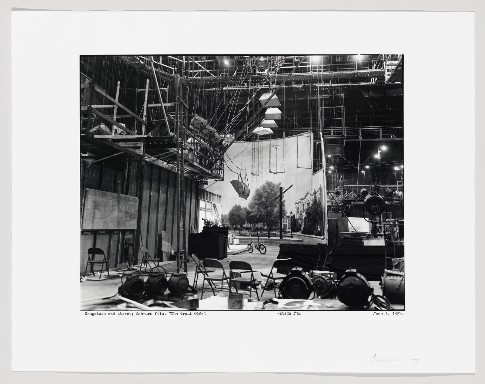 Black and white photograph of a film set for the feature film "The Great Gatsby," showing a constructed street scene with a drugstore facade, various stage equipment, and crew members working in the background. The image includes scaffolding, lighting rigs, and a bicycle in the foreground, with a caption at the bottom indicating the location as Stage #12 and the date as June 1, 1977.