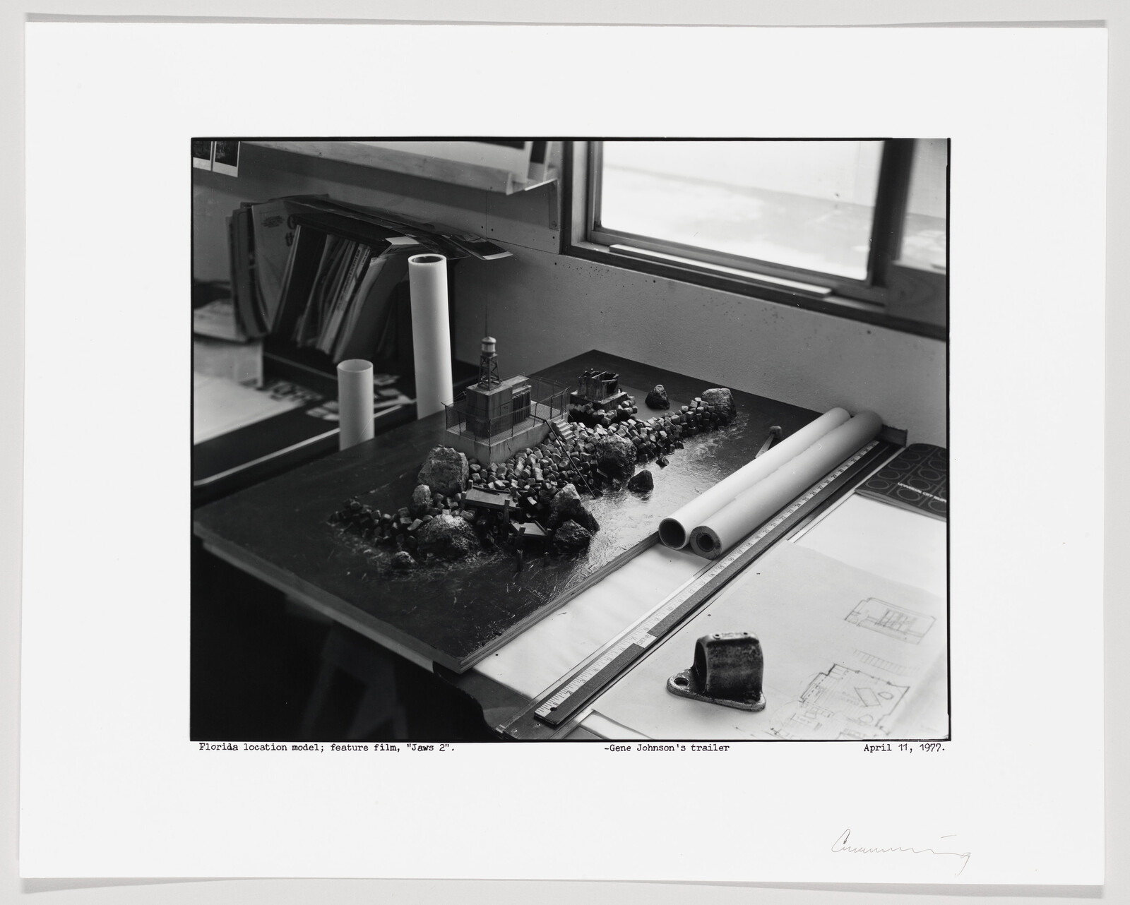 Detailed scale model of a rocky shoreline with buildings and drafting plans on a worktable.
