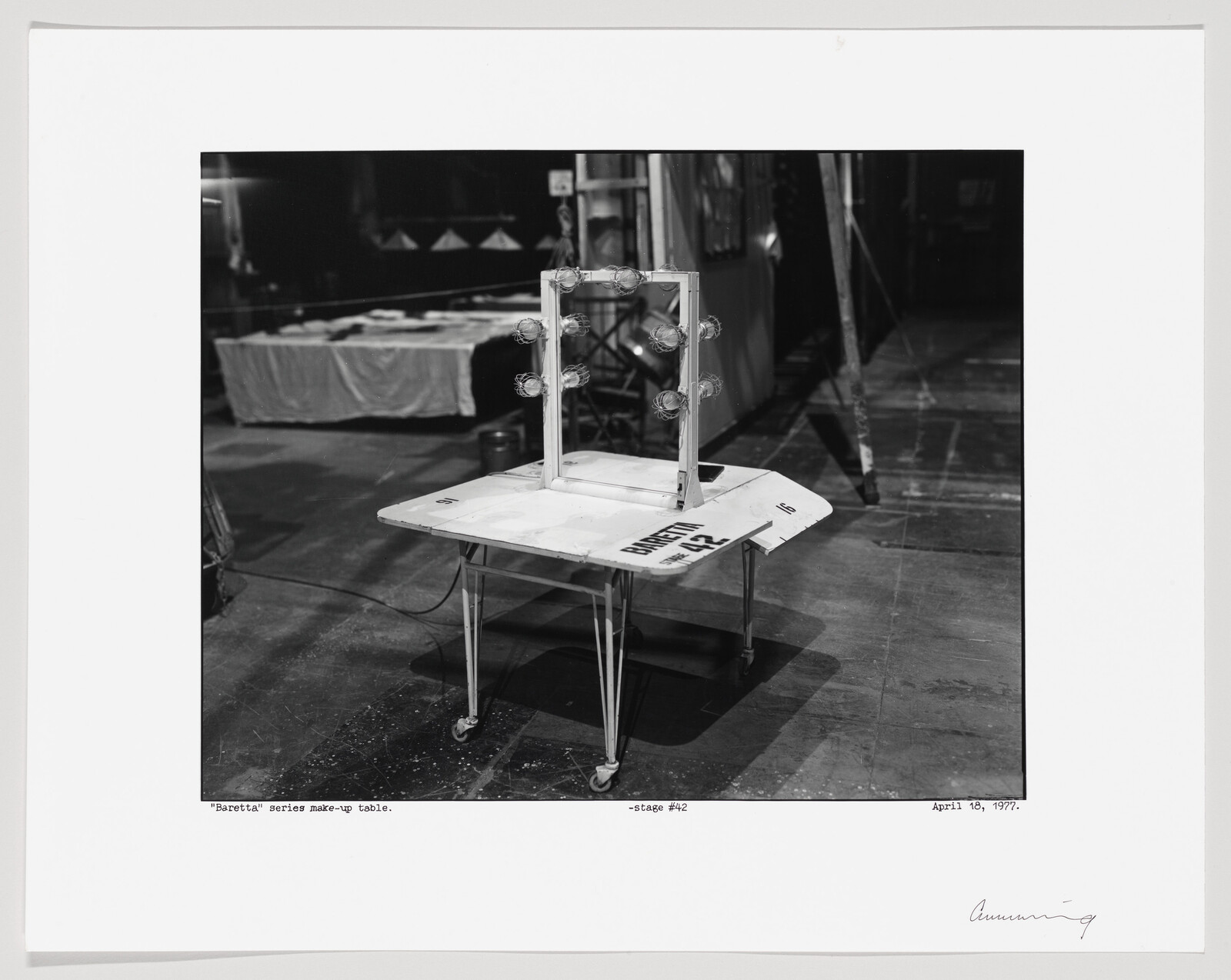 A black and white photograph depicting a makeup table with a clear, upright, rectangular panel with light bulbs around it, set on a wheeled table. The table is positioned in a backstage area with various items in the background, including a covered table and a ladder. The image includes handwritten notes and a date at the bottom, indicating the context as a makeup table from the "Barretta" series on stage #42, dated April 18, 1977.