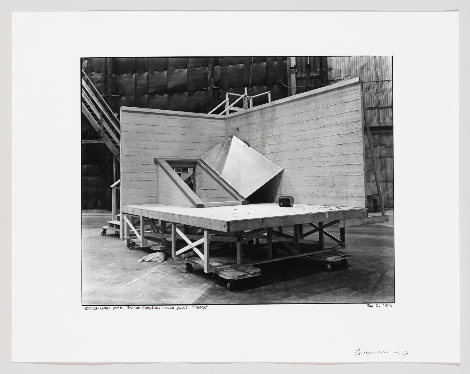 A wooden stage set with a tilted metal cube leaning against a cornered wall on casters.