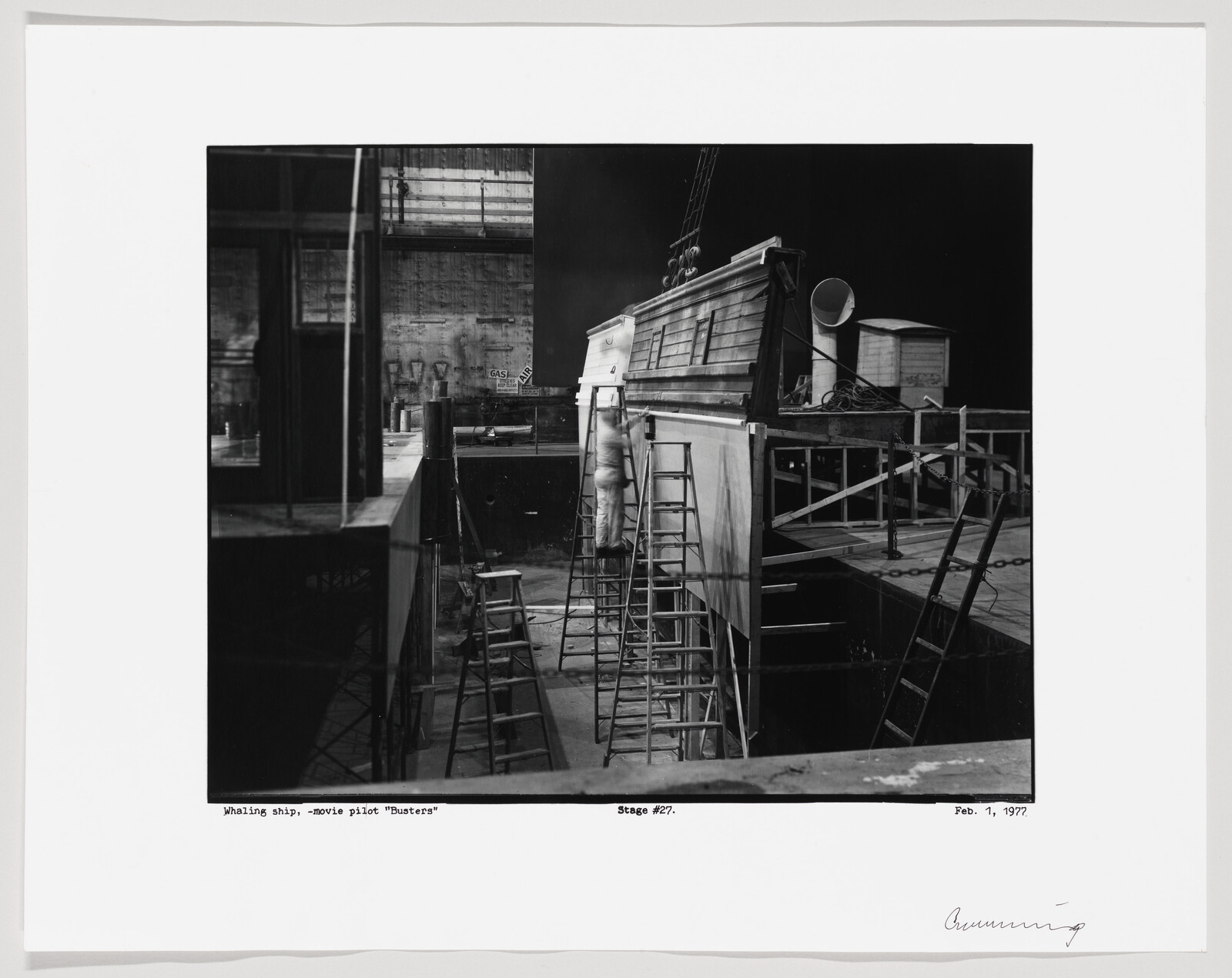 A worker climbs a ladder to work on a wooden ship set surrounded by scaffolding and ladders.