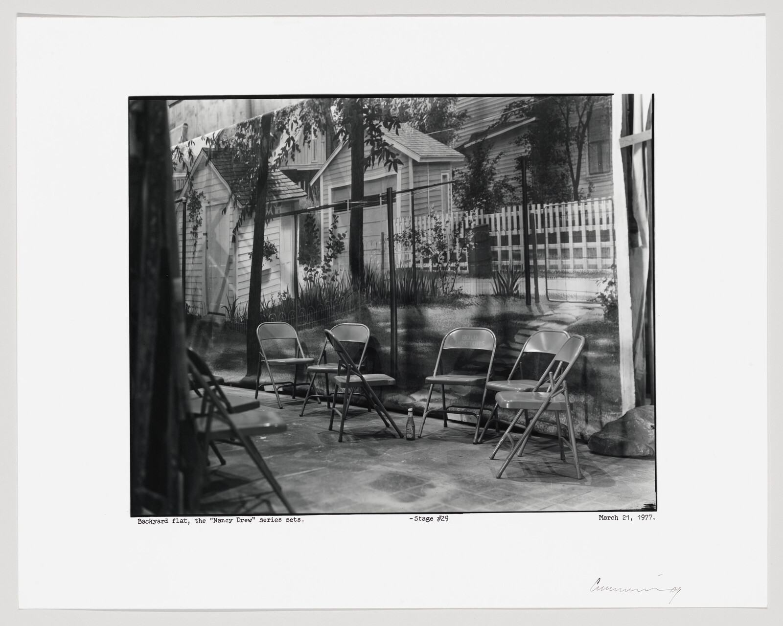A black and white photograph depicting a set of four metal folding chairs arranged on a concrete floor in front of a large backdrop with a printed image of a suburban backyard scene. The backdrop features a white picket fence, a house, and lush greenery. In the foreground, part of a dark structure is visible to the left, and a bottle sits on the ground near the chairs. The bottom of the image includes text that reads "Backyard flat, the 'Nancy Drew' series sets. -Stage #29 March 21, 1997," and there is a signature on the bottom right.