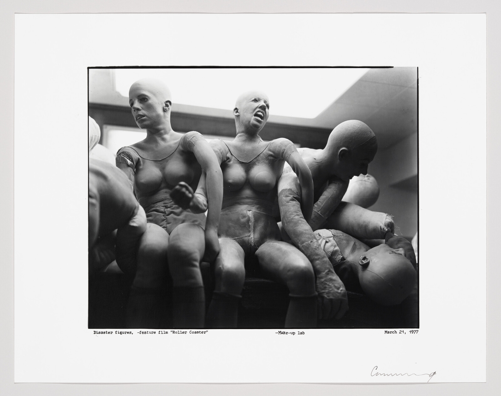 A black and white photograph showing a group of mannequins with exaggerated facial expressions and poses, suggesting panic or distress. They appear to be in a makeup lab, as indicated by the caption, and are possibly props for a feature film titled "Roller Coaster." The image is framed with a white border, and there is a handwritten signature at the bottom right corner.