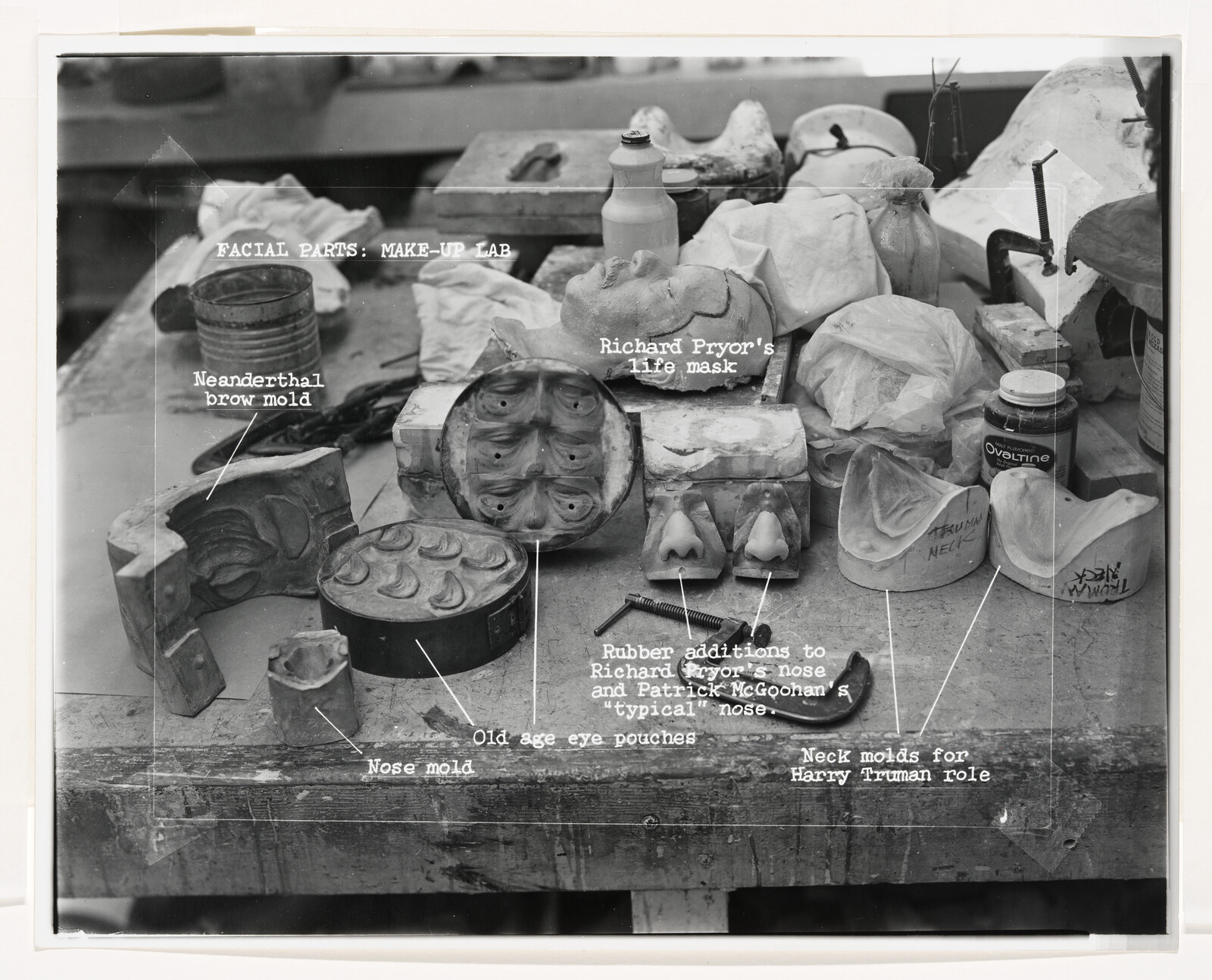 A cluttered makeup lab workbench with labeled facial molds, nose pieces, and a life mask.