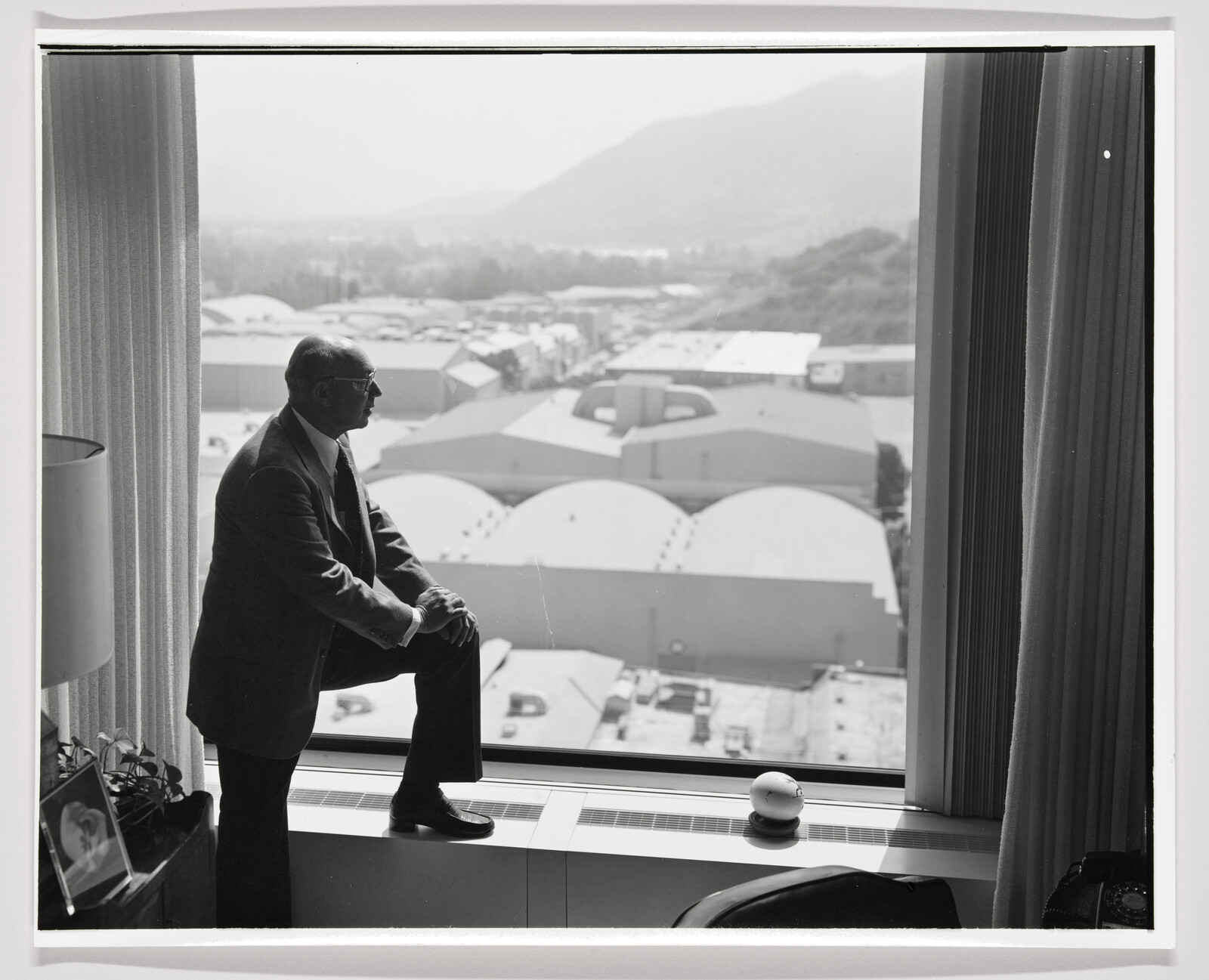 A black and white photo of a man in a suit and sunglasses sitting on a windowsill, looking out over industrial buildings with a mountainous backdrop. The interior includes a curtain partially drawn and various items on the windowsill, including a camera.
