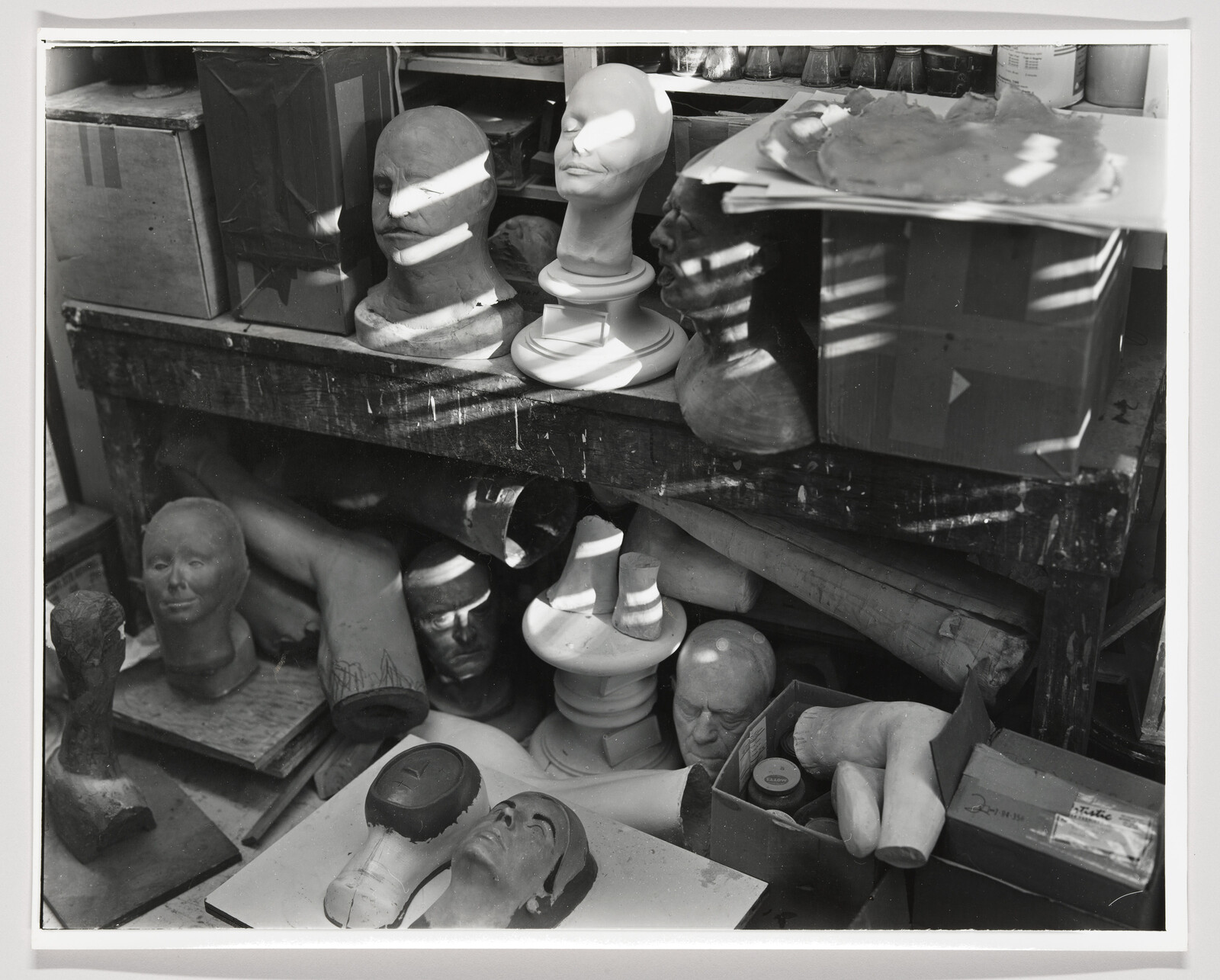 Several plaster mannequin heads and detached limbs clutter a workshop shelf under striped light.