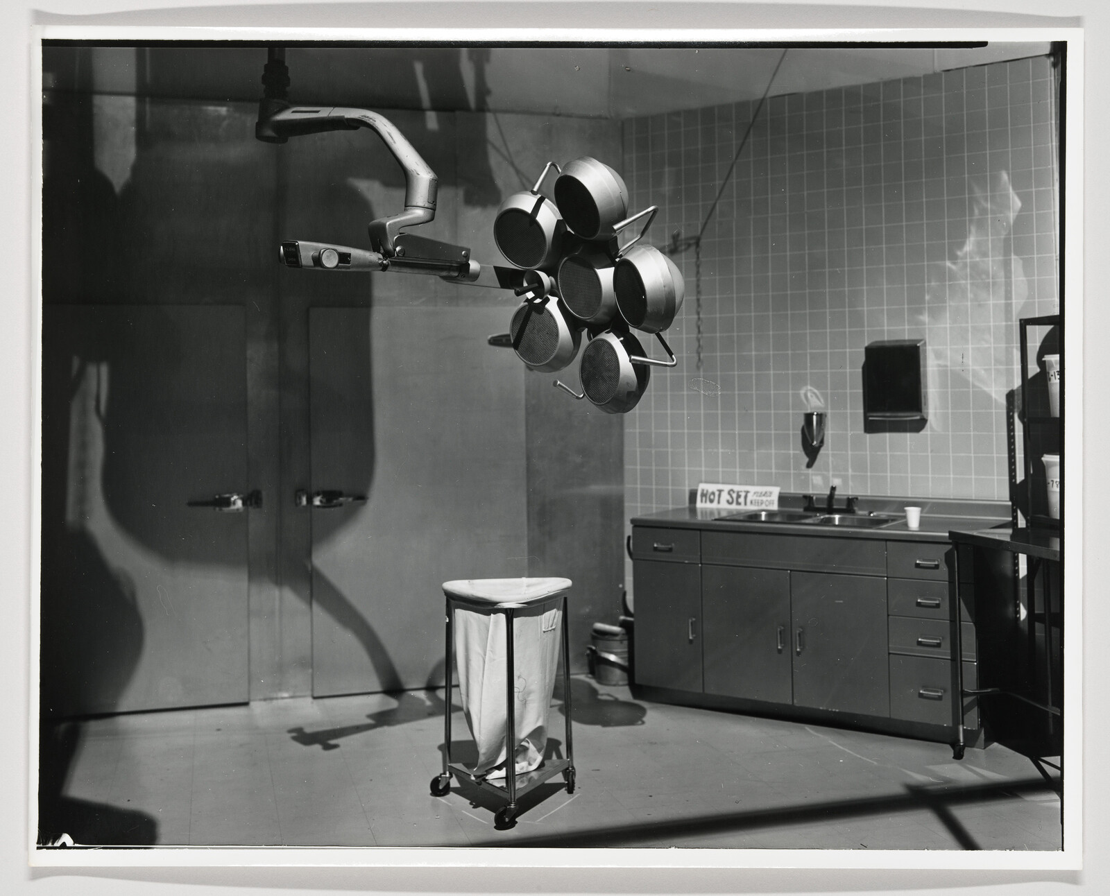 Overhead surgical lights hang above a wheeled linen cart in a tiled medical prep room.