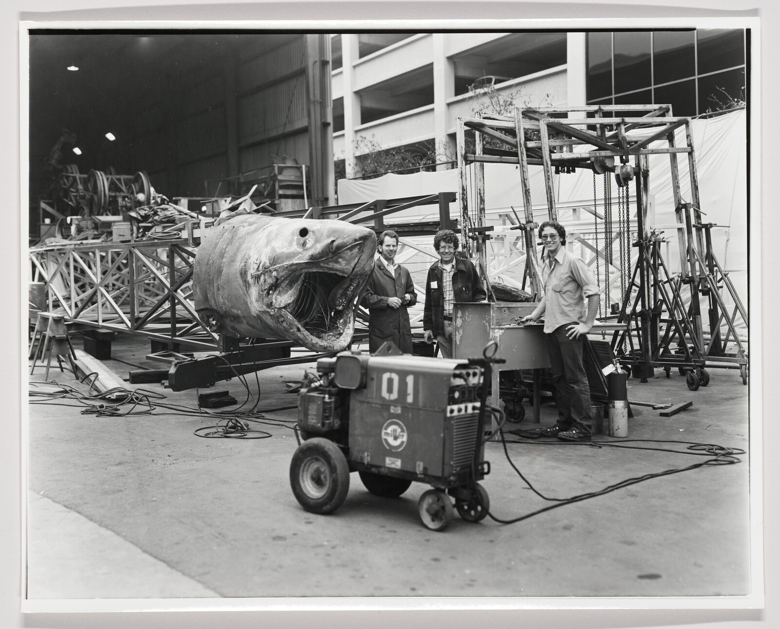 Three workers stand beside a large fabricated fish head and metal framework in a workshop.
