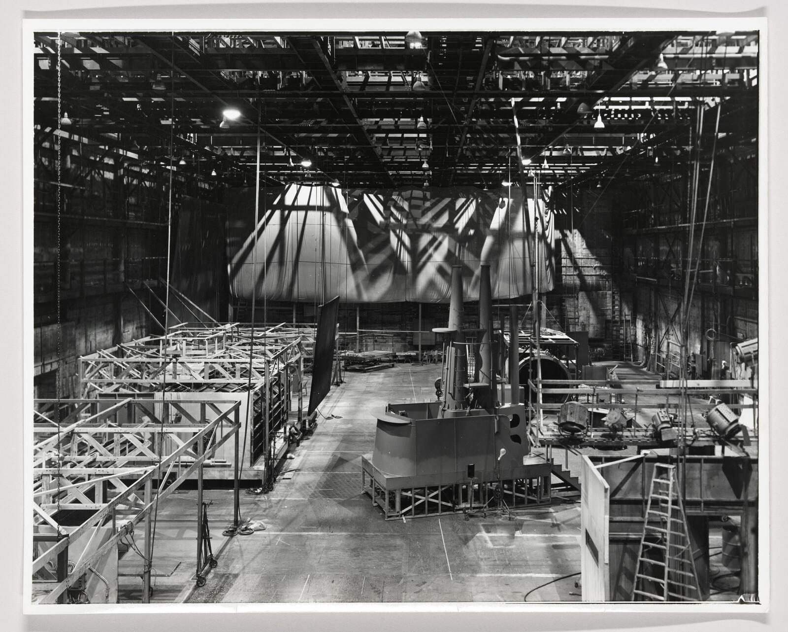 Large empty film soundstage with metal scaffolding, set structures, and overhead lighting rigs.