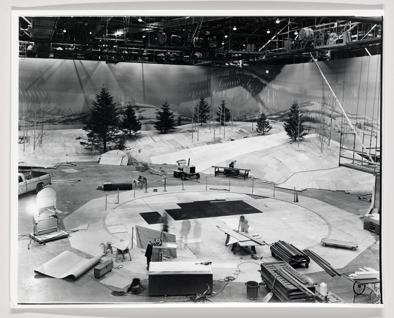 Film studio crew builds a snowy outdoor set with painted backdrop, trees, and scaffolding.