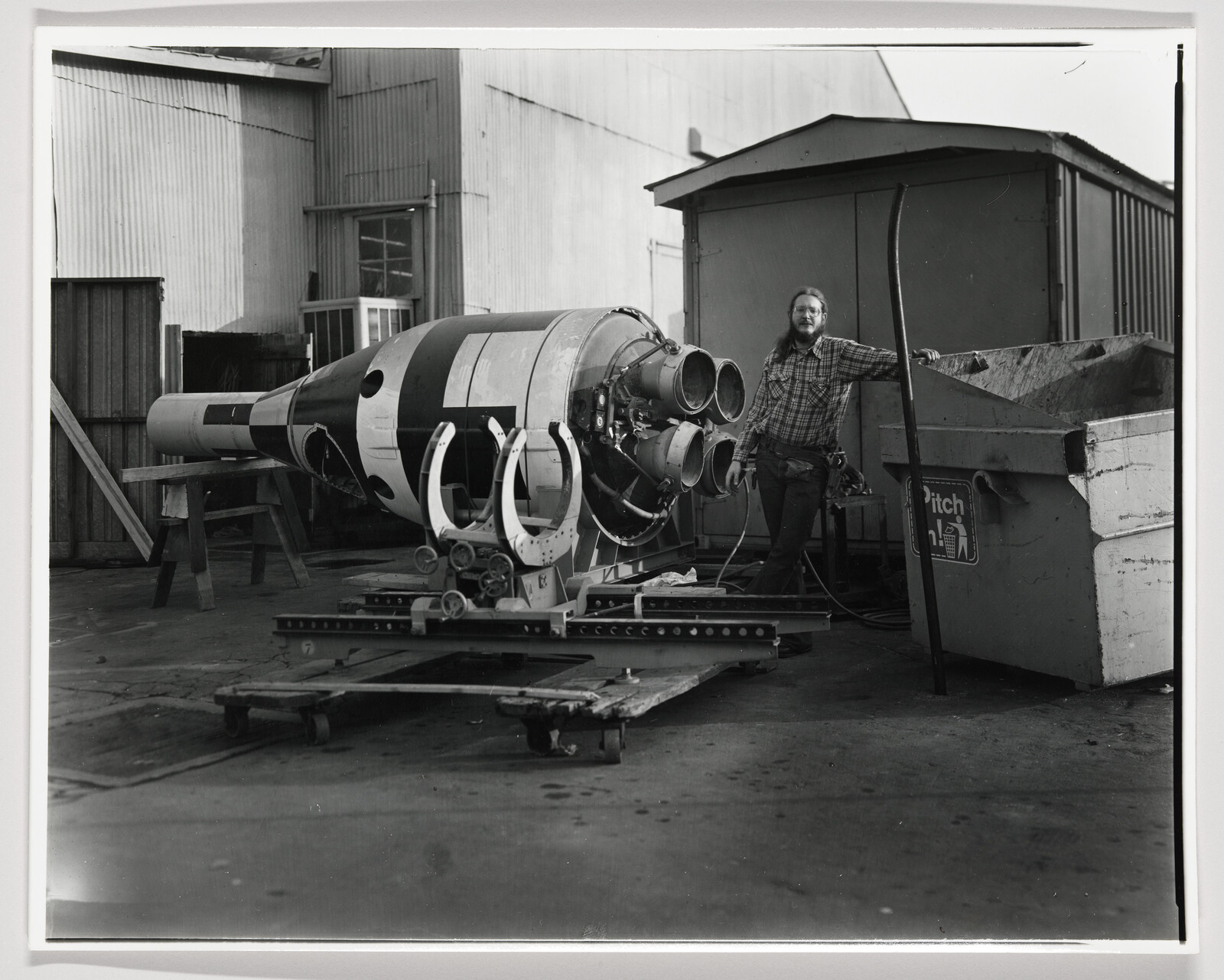 A man in a plaid shirt stands next to a large rocket engine section on a shop floor.