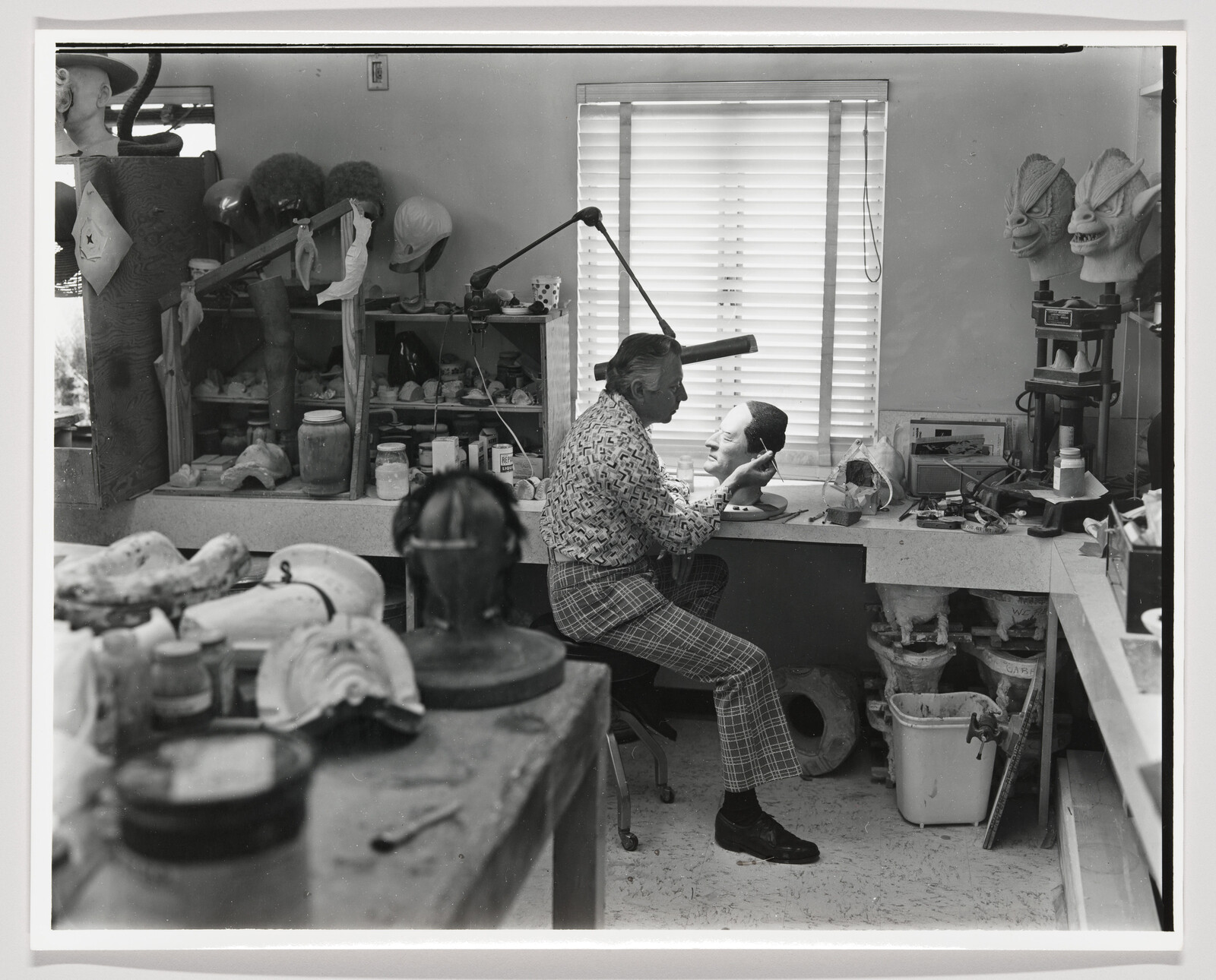 Man sculpts a lifelike head at a cluttered workbench in an effects studio.