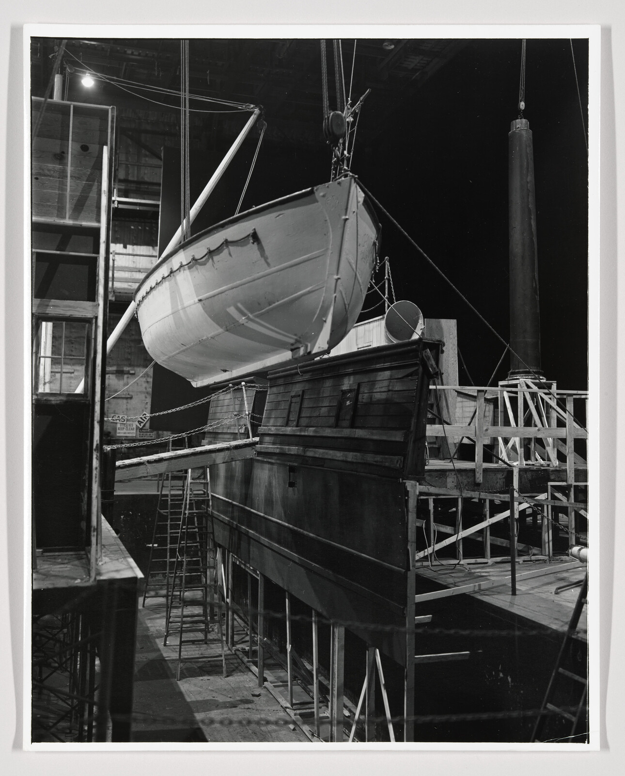 A lifeboat is suspended by cables above a wooden ship set while workers use ladders.