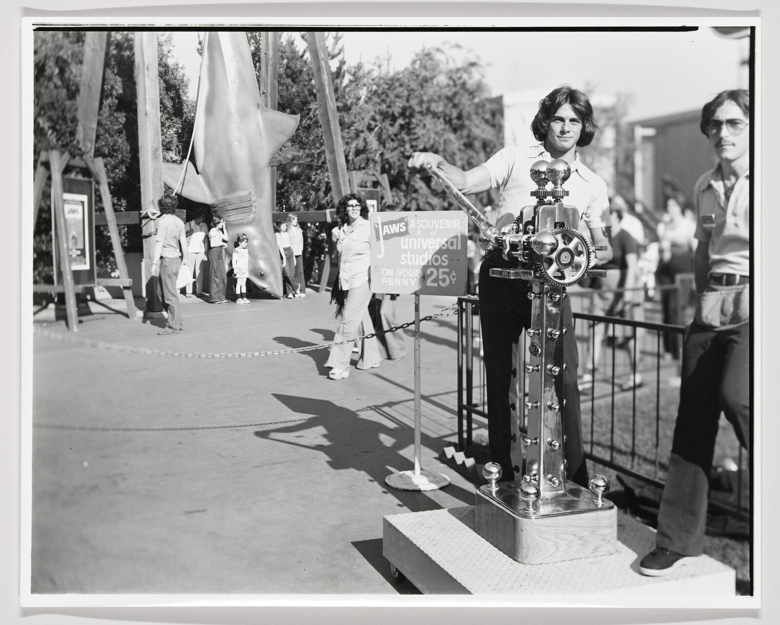 Young man cranks a penny-press souvenir machine while a large Jaws shark prop hangs nearby.