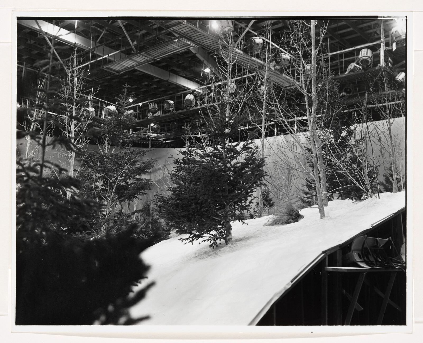 Studio set with snow-covered slope and evergreen trees under overhead lighting rigs.