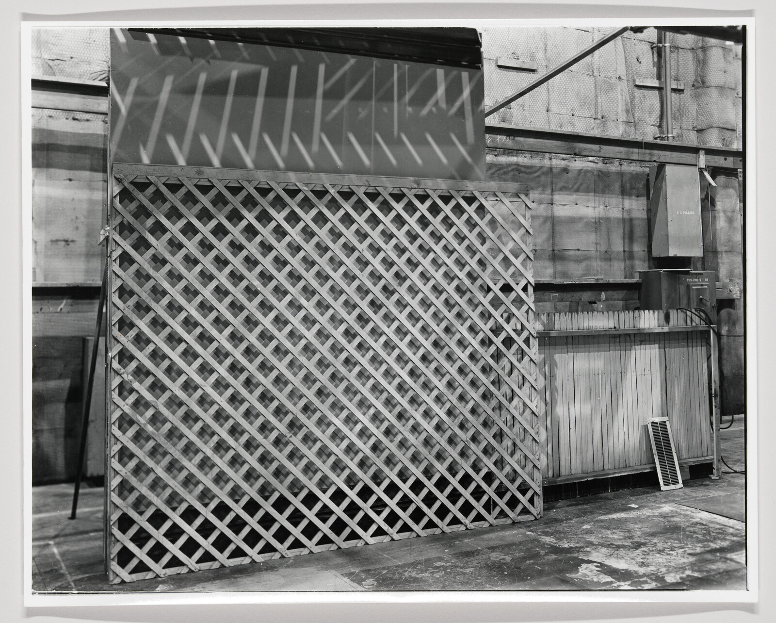 A black and white photograph showing an industrial setting with a large lattice structure in the foreground. To the right, there's a wooden wall with a metal box attached and a vent near the ground. The scene has a utilitarian appearance with shadows casting patterns on the lattice.