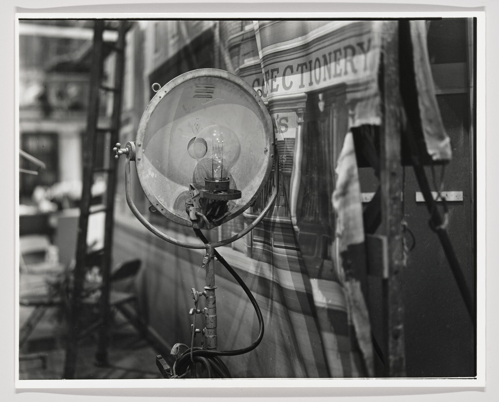 A vintage black and white photograph featuring a close-up of an old-fashioned, large round industrial light with a visible filament bulb, mounted on a metal stand. In the blurred background, part of a building with the word "CONFECTIONERY" on its facade is partially visible.