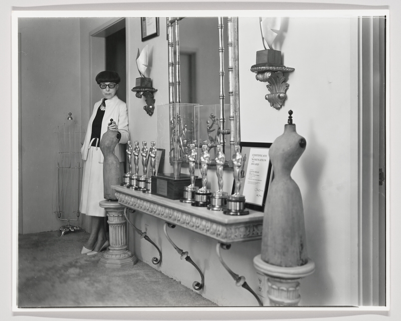 A black-and-white photograph of a woman standing in a room with a collection of trophies displayed on a shelf. She is wearing a white coat over a dark outfit, large round sunglasses, and is holding a trophy. To her left is a mannequin torso on a pedestal, and to her right is another mannequin torso, both without heads or limbs. The room has a vintage feel with ornate wall sconces and a framed certificate on the wall.