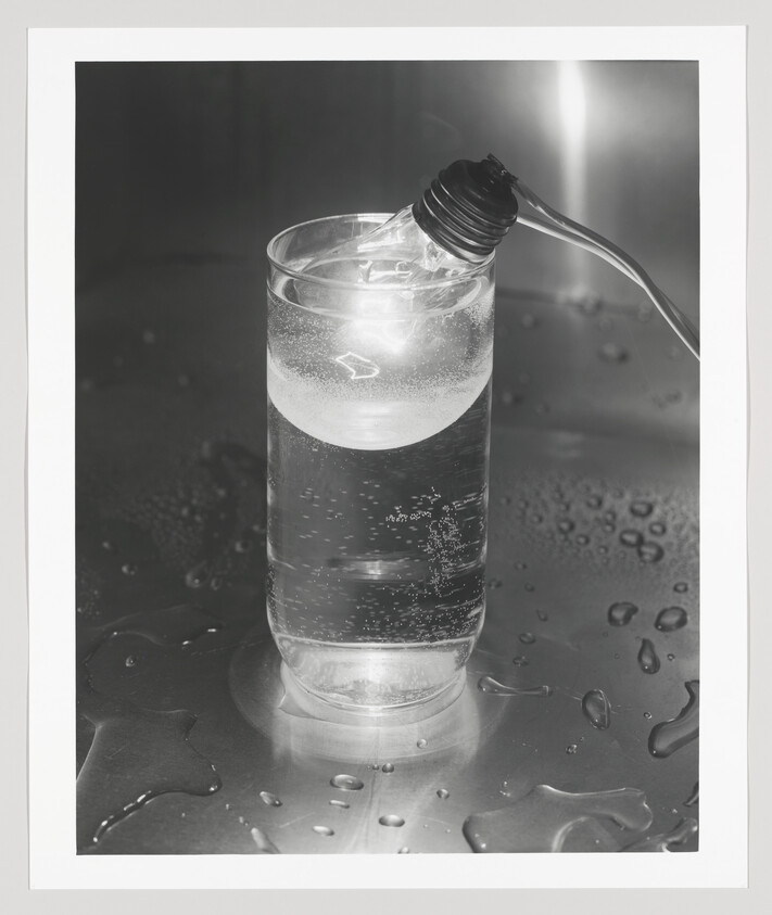 A lit light bulb rests on the rim of a glass filled with bubbling water on a wet surface.