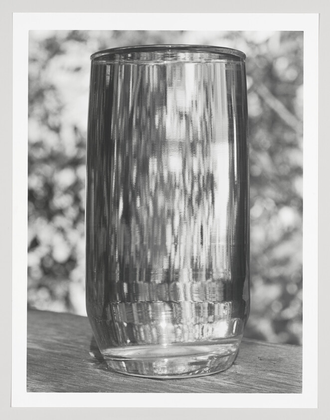 Tall clear drinking glass stands empty on a wooden surface with reflected light and blurred background.