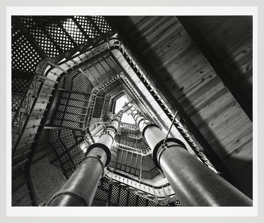 A black and white photograph looking up the interior of a spiral staircase with intricate metalwork and a wooden ceiling, framed by two large vertical pipes.