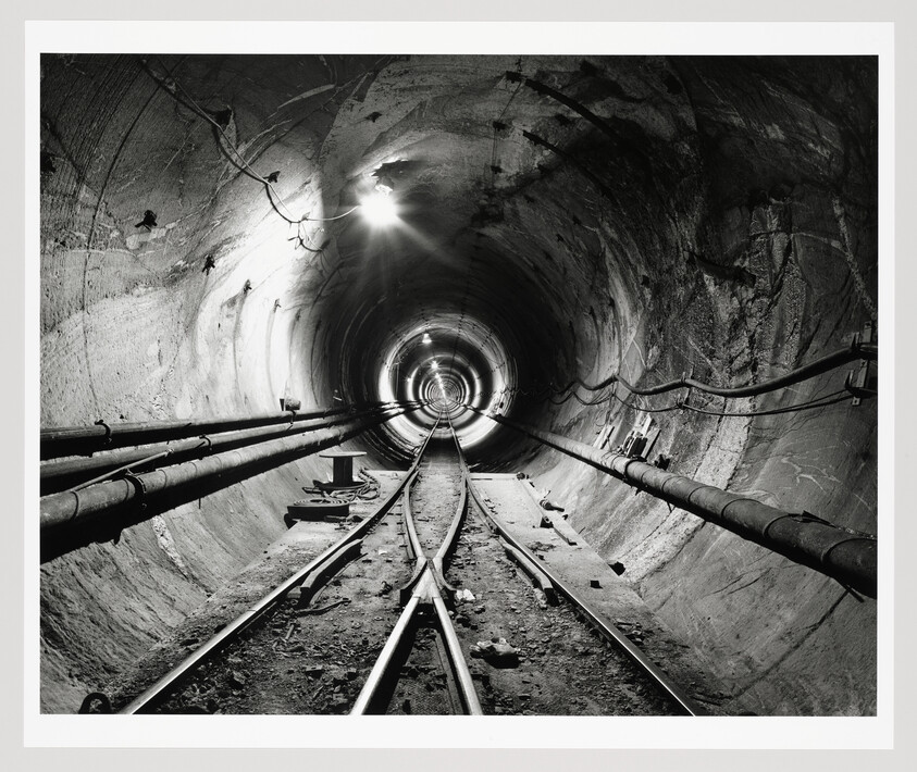 A black and white photograph of a railway tunnel with tracks leading into the distance. The tunnel is illuminated by a bright light at the far end and various smaller lights along the curved walls. Pipes and cables run alongside the tracks, and the rough texture of the tunnel walls is visible.