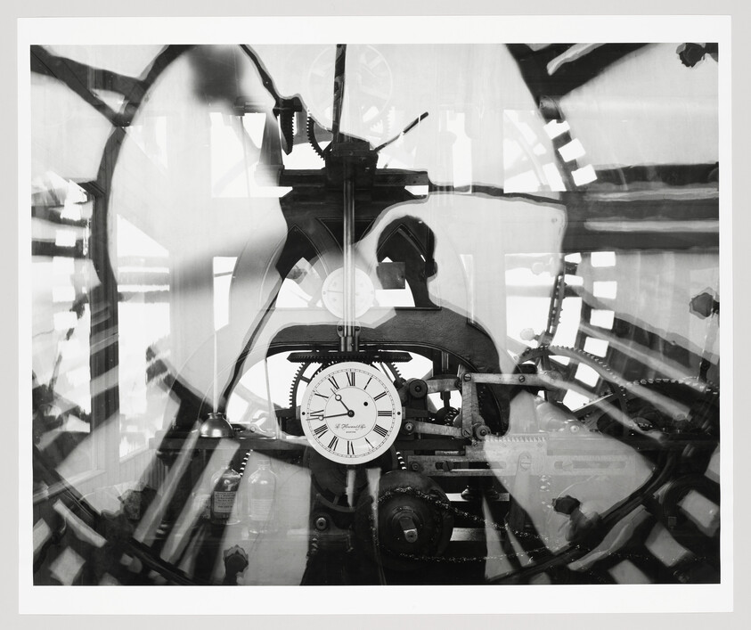 A black and white photograph showing the intricate inner workings of a large clock mechanism with visible gears, cogs, and a central clock face with Roman numerals. The image has a transparent, ghostly quality, suggesting motion or multiple exposures.