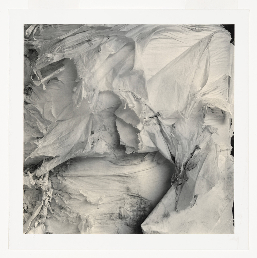 A black and white photograph depicting a close-up of a crumpled and torn piece of paper or fabric, with a variety of textures and shadows creating an abstract composition. The image is bordered by a white frame, suggesting it may be a print or a photograph of an artwork.