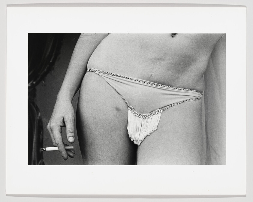 Woman wearing fringed bikini bottom holds a lit cigarette near her hip.