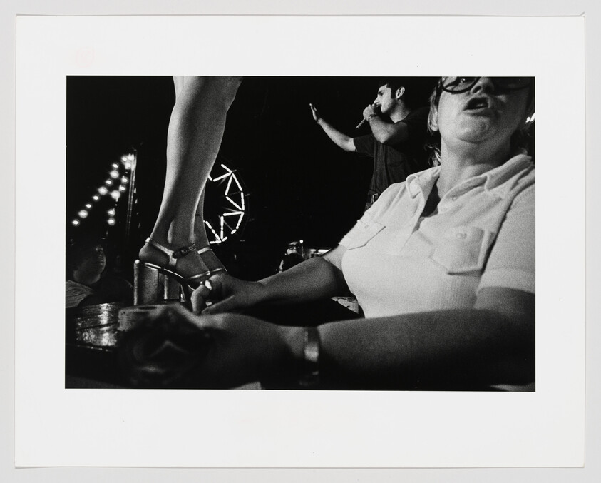 A black and white photograph capturing a lively scene, possibly at a concert or public event. In the foreground, a woman is seated with her arms crossed, wearing glasses and a collared shirt. To the left, another person's legs are visible, standing with one foot on a raised surface, wearing high-heeled sandals. In the background, a man is holding a microphone, suggesting he is performing or speaking to the crowd. Lights are strung up in the distance, contributing to the festive atmosphere.