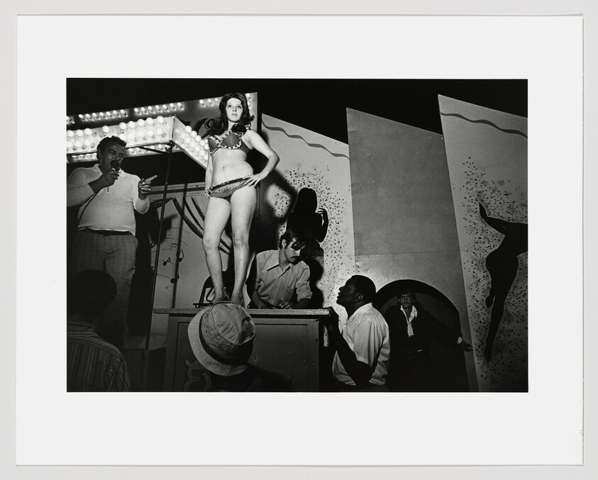 A woman in a bikini stands on a stage platform while men watch and speak into a microphone.