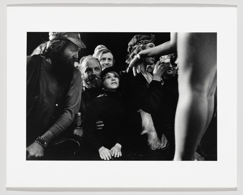 A black and white photograph capturing a group of people looking intently towards the right, with a partial view of a nude figure in the foreground on the right side. The group appears engaged and focused, with various expressions of curiosity and interest. The setting seems informal, possibly at an event or gathering.
