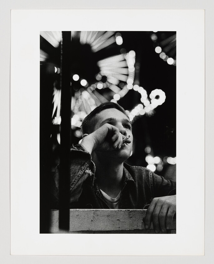 A young man leans on a railing and smokes while carnival lights glow behind him.