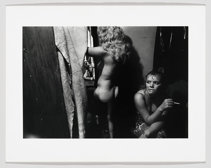 Two young women in a dim dressing room, one nude adjusting a curtain while the other smokes.